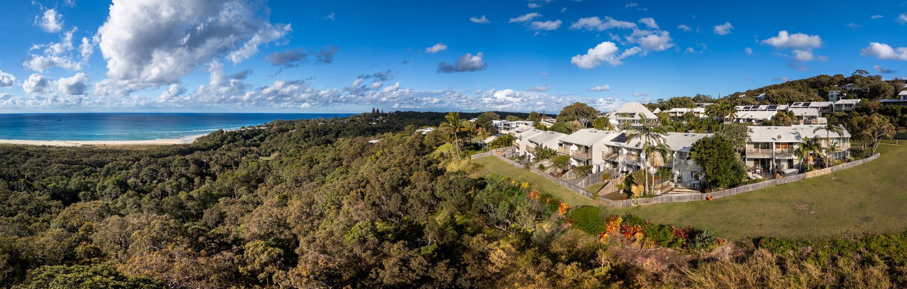 Panorama over the coastal bushland below Pandanus Palms Resort to the long surf beach and the ocean, with the villas on the headland at right