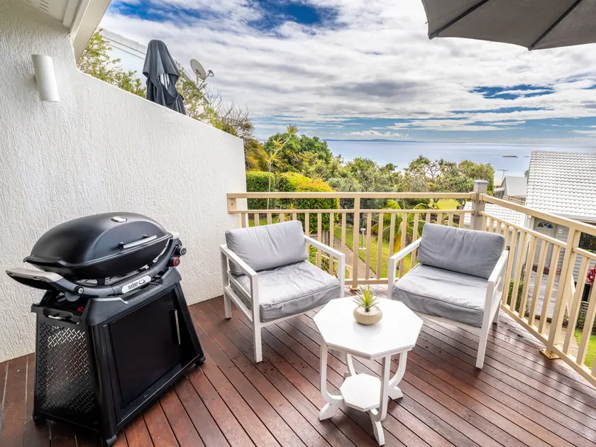 A villa balcony with a barbecue and two armchairs, looking over the treetops to the ocean