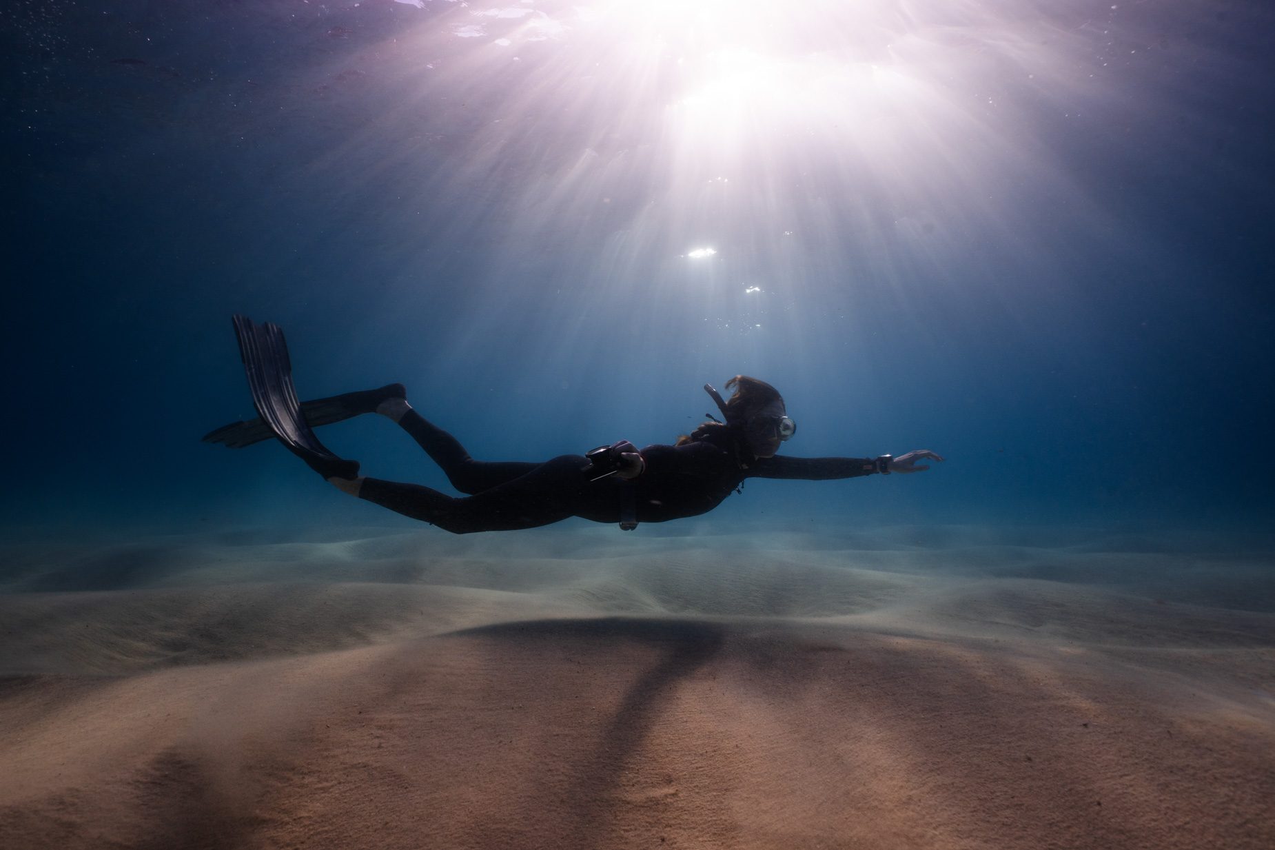 A freediver gliding over the sandy ocean floor as sunlight streams through the water