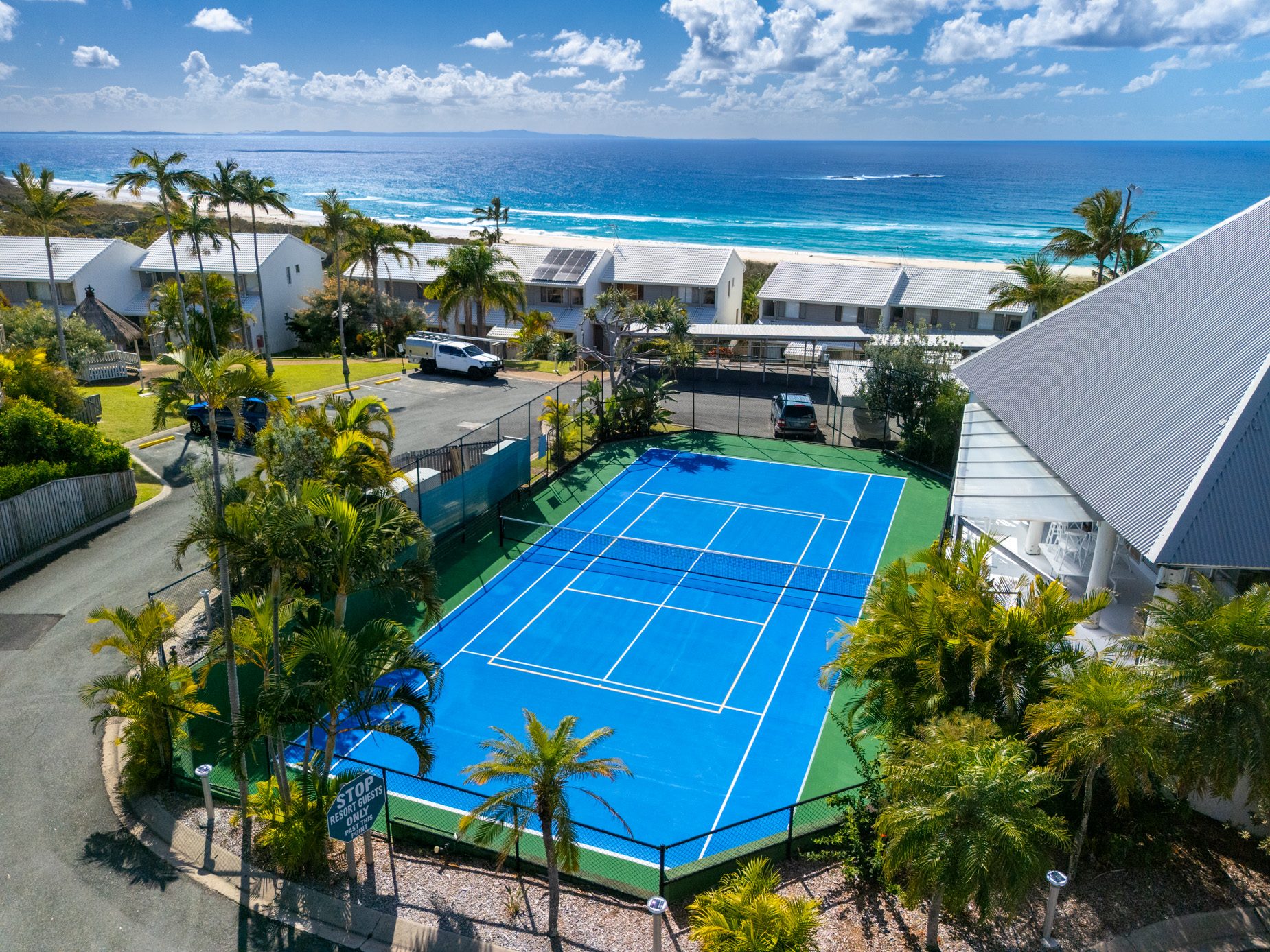 Pandanus Palms Resort from the air: a row of villas above the beach and the ocean, with the tennis and pickle ball court in the foreground